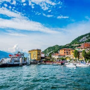 A ferry boat is docked at a pier in a colorful lakeside town, with lush green mountains rising behind it under a blue, cloudy sky.