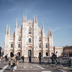 An ornate, white Gothic cathedral stands behind a bustling public square filled with people on a sunny day.