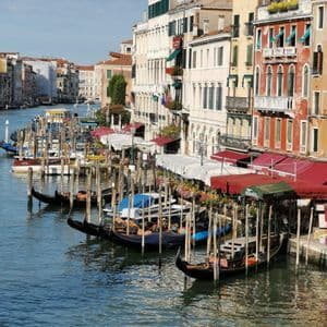 A row of gondolas is moored to wooden posts in a canal, with colorful, multi-story buildings lining the waterway under a blue sky.