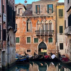 Gondolas are docked in a narrow canal, with their reflections mirrored in the water beside weathered brick buildings.