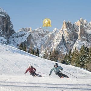 Due persone sciano su un'ampia pista battuta, con uno sfondo di montagne innevate sotto un cielo azzurro e limpido.