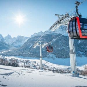 Funivie rosse attraversano un vasto paesaggio innevato, con una valle e montagne illuminate dal sole sullo sfondo.