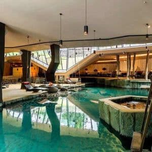 L'interno di una spa moderna con una grande piscina turchese, altalene sospese, aree lounge e grandi pilastri strutturali.