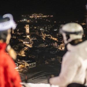 Un viaggio di gruppo WeRoad di sciatori su una pista di notte, che guarda giù un villaggio di montagna sottostante illuminato a festa.