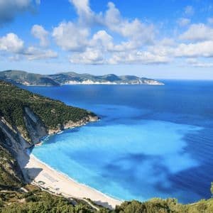 Vista dall'alto di una spiaggia di sabbia bianca e di un mare turchese in una cala, incorniciata da ripide scogliere verdi sotto un cielo parzialmente nuvoloso.