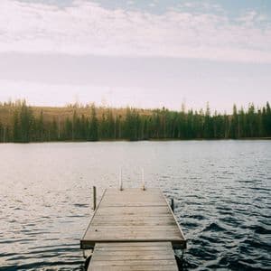 Un ponton en bois avec une petite échelle s'avance dans un lac, une forêt dense bordant la rive lointaine sous un ciel partiellement nuageux.