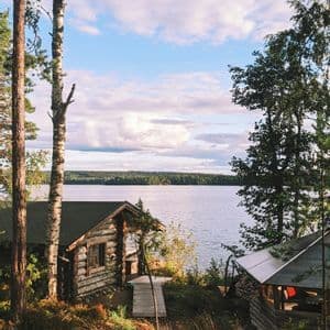 Une cabane en rondins rustique se dresse sur une berge herbeuse, dominant un grand lac, avec une forêt dense sur la rive opposée sous un ciel nuageux.