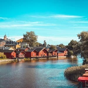 Une rangée de maisons en bois rouges borde la rive d'une rivière sous un ciel bleu clair, avec des arbres en arrière-plan.