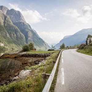 Una carretera pavimentada serpentea a lo largo de un fiordo, con empinadas montañas verdes emergiendo del agua bajo un cielo parcialmente nublado.
