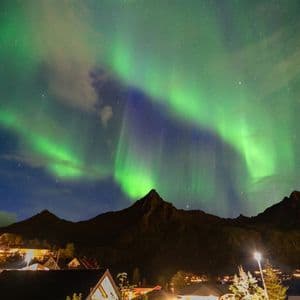 La aurora boreal resplandece en verde y púrpura en un cielo nocturno estrellado sobre la silueta oscura de montañas y un pueblo iluminado.