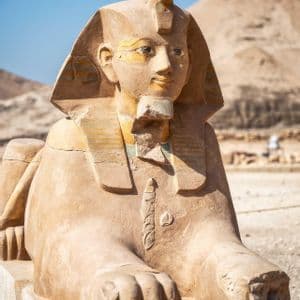 Eine antike Sandstein-Sphinx-Statue ruht auf einem Sockel bei hellem Tageslicht in einer Wüstenlandschaft.