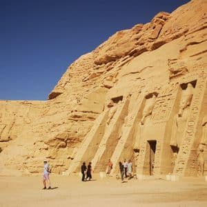 Touristen gehen auf sandigem Boden vor kolossalen antiken Statuen, die in eine Sandsteinklippe gehauen sind, unter einem klaren blauen Himmel.