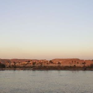 Ein breiter Fluss mit Palmen und einem kleinen Dorf am Ufer vor trockenen Hügeln unter einem klaren, blassen Himmel.
