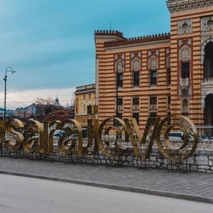 Un grande cartello dorato con la scritta 'Sarajevo' su un marciapiede di fronte a un edificio ornato e a strisce sotto un cielo nuvoloso.