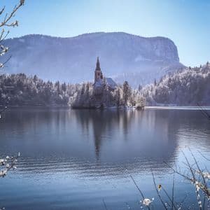 A church on a snow-covered island in a lake, surrounded by frosty trees and distant mountains under a clear blue sky.