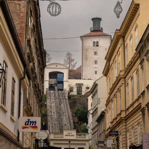 A blue funicular climbs a steep track on a narrow city street, with a historic tower visible in the background.