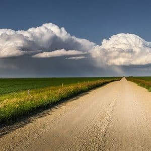 Una lunga strada sterrata si estende attraverso ampi campi verdi sotto una grande nuvola temporalesca bianca contro un cielo azzurro.