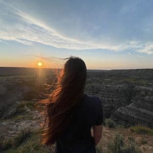 Una donna vista di spalle, con lunghi capelli al vento, ammira un vasto paesaggio di canyon al tramonto.