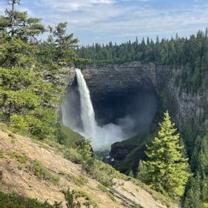 Una potente cascata si getta in un profondo canyon circolare, circondato da una fitta foresta sempreverde, sotto un cielo parzialmente nuvoloso.