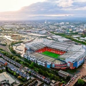 Una veduta aerea di un grande stadio di calcio con spalti rossi e un campo verde circondato da un paesaggio urbano.
