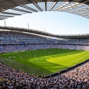 Gli spettatori riempiono un grande stadio che circonda un campo da calcio verde brillante durante una partita diurna.