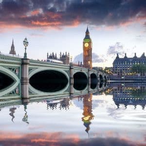Un ponte ad arco attraversa un fiume verso una torre dell'orologio illuminata, con nuvole colorate del tramonto che si riflettono sull'acqua.