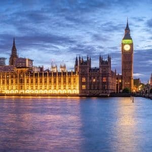 Un parlamento gotico illuminato e una torre dell'orologio accanto a un ampio fiume al crepuscolo con un ponte ad arco