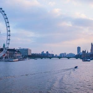 Una grande ruota panoramica e il paesaggio urbano lungo un ampio fiume con barche che navigano sotto un cielo crepuscolare.