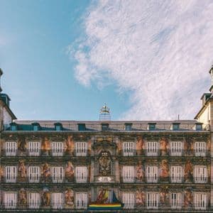 Facciata di edificio storico con elaborati affreschi murali, balconi multipli e torri dell'orologio a doppia guglia sotto un cielo azzurro.