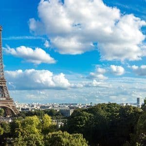 Un'alta torre a traliccio si erge sopra un paesaggio urbano circondato da alberi verdi sotto un cielo azzurro luminoso con nuvole bianche.