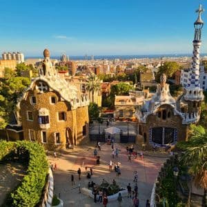 Visitantes caminando por ornamentados pabellones de piedra con agujas de mosaico con vistas a la ciudad y al mar bajo un cielo despejado.