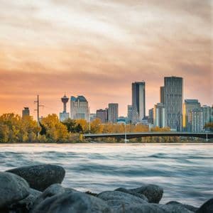 A city skyline rises behind a flowing river and a rocky bank under a cloudy sunset sky.