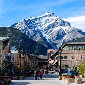 People walk down a paved pedestrian street in a town against the backdrop of a large snow-covered mountain peak.