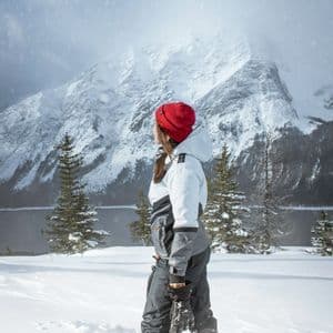 A person in winter clothing and a red beanie stands in deep snow overlooking a lake and snow-covered mountain.