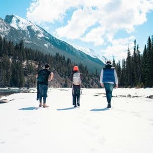A WeRoad group trip of three people walking on snow along a river valley framed by pine trees and mountains.