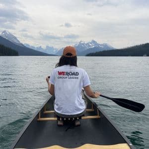 Un coordinatore di WeRoad pagaia in kanoa attraverso un ampio lago circondato da rive bosco-secche e cime montuose.