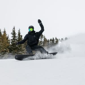 Un snowboarder vestido de negro descendiendo por una pendiente nevada, levantando nieve polvo fresca con pinos al fondo.