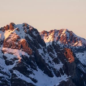 Picos de montañas rocosas cubiertas de nieve iluminados por una cálida luz solar dorada contra un cielo pálido.