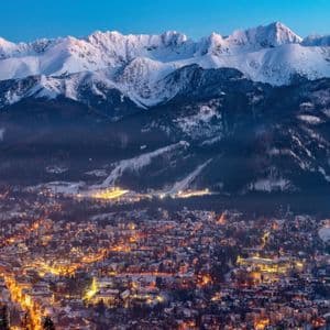 An illuminated town glows at dusk beneath towering snow-covered mountain peaks and forested slopes.