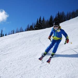 A skier wearing a helmet and blue ski suit skiing down a snowy mountain slope under a clear blue sky.