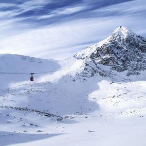 A red cable car travels across a snow-covered mountain valley under a partly cloudy blue sky.
