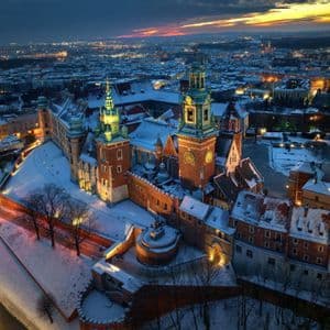 An illuminated historic brick castle with towers stands covered in snow overlooking a city at dusk.