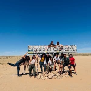 Un viaggio di gruppo WeRoad in posa vicino al cartello del Tropico del Capricorno in un deserto arido sotto un cielo azzurro