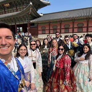 Un viaje en grupo WeRoad, vistiendo ropa tradicional coreana, posa para una selfie grupal frente a un edificio histórico.