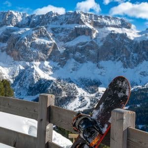 Uno snowboard è appoggiato a una ringhiera di legno che si affaccia su montagne aspre e innevate sotto un cielo azzurro.
