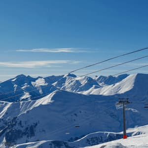 Sciatori su una cresta di montagna innevata con seggiovie e cavi di telecabina contro cime innevate sotto un cielo azzurro.
