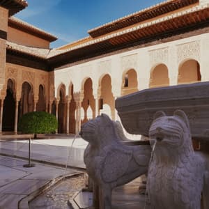 Una fuente de mármol con estatuas de leones arrojando agua en un patio soleado rodeado de intrincadas columnatas arqueadas.