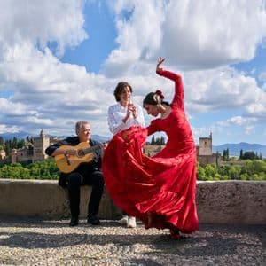 Una mujer con vestido rojo baila flamenco junto a un guitarrista y una mujer tocando las palmas al aire libre con vistas a una fortaleza histórica.