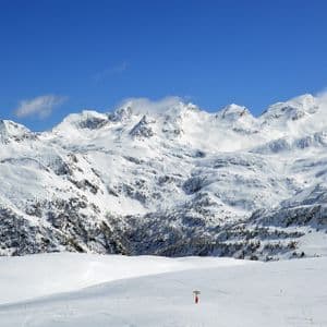 Cime montuose innevate si ergono dietro a una vasta pista da sci bianca sotto un cielo azzurro e limpido.