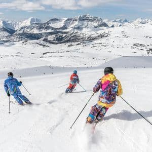 Eine WeRoad-Gruppenreise von Skifahrern in bunten Anzügen, die einen schneebedeckten Hang mit Blick auf riesige Gipfel hinabfahren